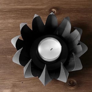 May include: A black metal candle holder shaped like a flower, with a white candle in the center. The petals have a folded design, and the holder sits on a wooden surface. The candle holder is a decorative item.
