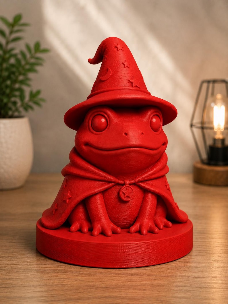 May include: A red frog figurine wearing a wizard hat and cape. The hat has star and crescent moon details. The frog is sitting on a round base. The cape is fastened with a star-shaped clasp. The figurine is a whimsical decorative item.