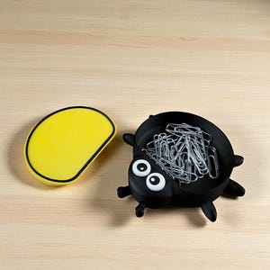 Puede incluir: Un organizador de escritorio negro con forma de bicho lleno de clips plateados, junto a un recipiente amarillo de forma ovalada. El organizador tiene ojos blancos y peque&ntilde;as patas negras. Ambos art&iacute;culos est&aacute;n sobre una superficie de madera clara.