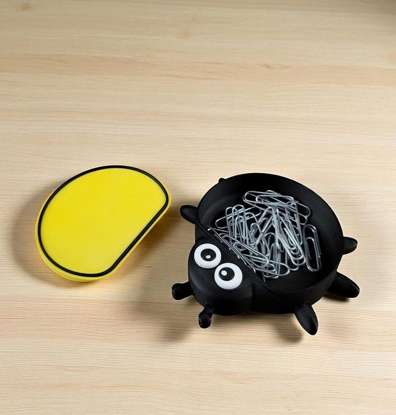 Puede incluir: Un organizador de escritorio negro con forma de bicho lleno de clips plateados, junto a un recipiente amarillo de forma ovalada. El organizador tiene ojos blancos y peque&ntilde;as patas negras. Ambos art&iacute;culos est&aacute;n sobre una superficie de madera clara.