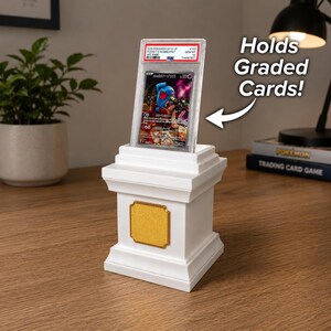 May include: A white display stand with a gold accent holds a graded Pok&eacute;mon card. The stand has a rectangular gold plaque and a tiered design. The text "Holds Graded Cards!" is next to the stand.