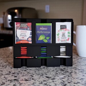 May include: A black tea organizer with three sections for tea bags and three lower compartments for individual tea packets. The tea bags are labeled "King's Crown Winter Punch", "Me&szlig;mer Pfefferminze", and "King's Crown Zimt Apfel". A white mug is in the background.