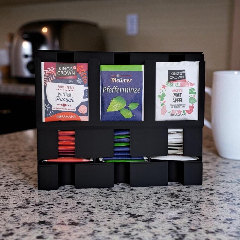 May include: A black tea organizer with three sections for tea bags and three lower compartments for individual tea packets. The tea bags are labeled "King's Crown Winter Punch", "Me&szlig;mer Pfefferminze", and "King's Crown Zimt Apfel". A white mug is in the background.