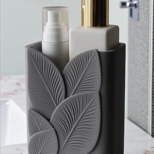 May include: A gray bathroom organizer with a leaf design, holding a white lotion bottle and a white and gold soap dispenser. The organizer is on a marble surface.