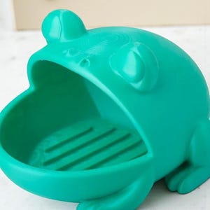 May include: A teal frog-shaped soap dish. The dish has a wide open mouth and a textured interior. The frog has large eyes and a detailed design. The dish is made of a solid material and is designed to hold soap.