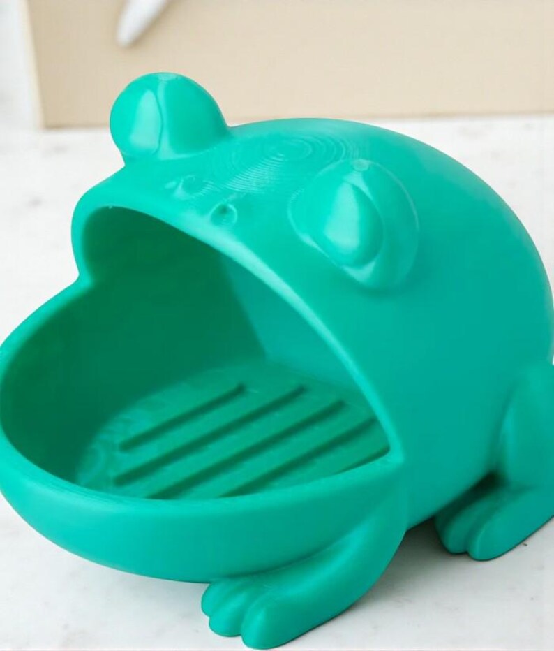 May include: A teal frog-shaped soap dish. The dish has a wide open mouth and a textured interior. The frog has large eyes and a detailed design. The dish is made of a solid material and is designed to hold soap.