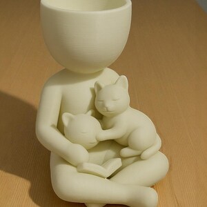 May include: A cream-coloured planter in the form of a person cradling two sleeping cats. The person is seated with a book, and the planter's head is a large, open bowl. The cats are nestled in the person's arms.