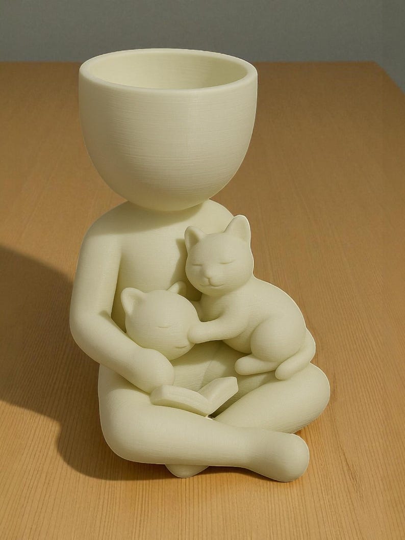 May include: A cream-coloured planter in the form of a person cradling two sleeping cats. The person is seated with a book, and the planter's head is a large, open bowl. The cats are nestled in the person's arms.