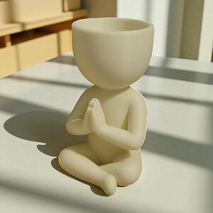 Puede incluir: Una maceta con forma humana de color crema, con las manos juntas en oraci&oacute;n. La maceta tiene una cabeza redondeada con una abertura profunda para las plantas. La figura est&aacute; sentada con las piernas cruzadas. El dise&ntilde;o general es minimalista y moderno.