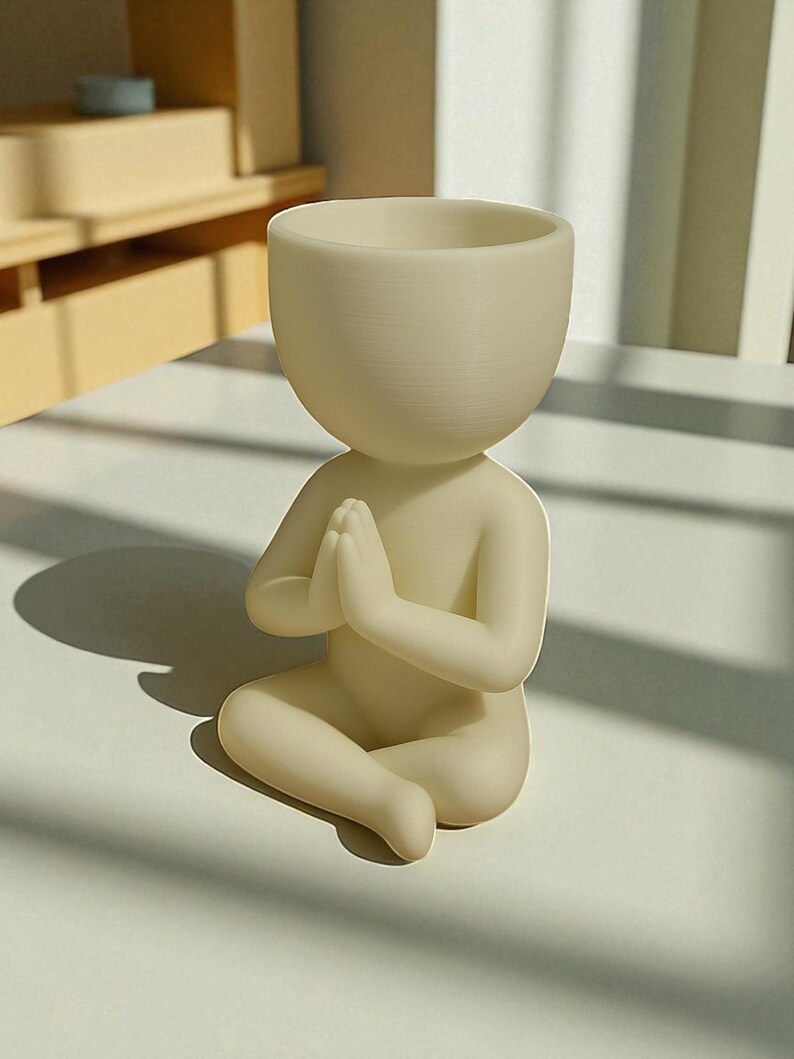 Puede incluir: Una maceta con forma humana de color crema, con las manos juntas en oraci&oacute;n. La maceta tiene una cabeza redondeada con una abertura profunda para las plantas. La figura est&aacute; sentada con las piernas cruzadas. El dise&ntilde;o general es minimalista y moderno.