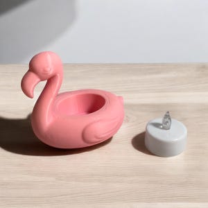 May include: A pink flamingo-shaped candle holder with a round opening for a candle. A small, white, battery-operated LED candle is next to the flamingo. The candle has a clear, flame-shaped top. The items are on a light wood surface.