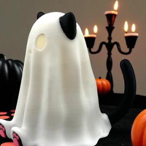 May include: A white Halloween decoration shaped like a ghost cat. The cat has black ears and a black tail. The ghost cat has pink paw pads. A black candelabra and pumpkins are in the background.