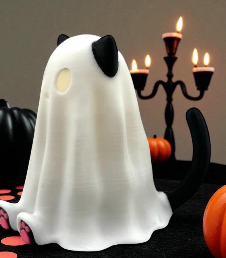 May include: A white Halloween decoration shaped like a ghost cat. The cat has black ears and a black tail. The ghost cat has pink paw pads. A black candelabra and pumpkins are in the background.