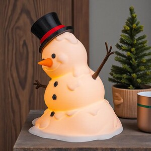 May include: A novelty snowman lamp with a melting effect. It has a black top hat with a red band, a carrot nose, and twig arms. The snowman has three black button details and gives off a warm glow. A festive Christmas decoration for the home.