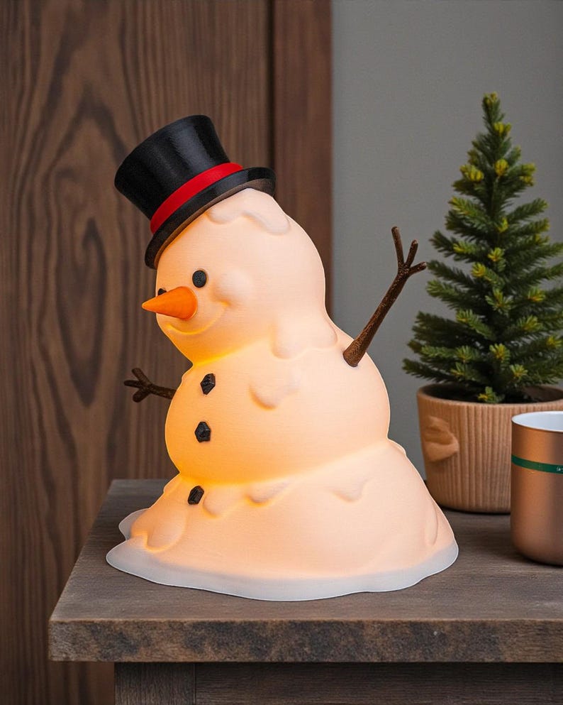 May include: A novelty snowman lamp with a melting effect. It has a black top hat with a red band, a carrot nose, and twig arms. The snowman has three black button details and gives off a warm glow. A festive Christmas decoration for the home.