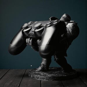 May include: A black and gray sculpture of a knight holding a large video game controller. The knight is in a stooped position, with the controller prominently displayed. The sculpture is on a decorative base, set against a dark background.