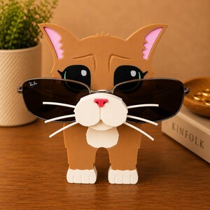 May include: A brown cat-shaped sunglasses holder with black sunglasses. The holder has pink inner ears, a white muzzle, and white whiskers. The sunglasses have a silver frame and dark lenses. The cat is standing on a wooden surface.