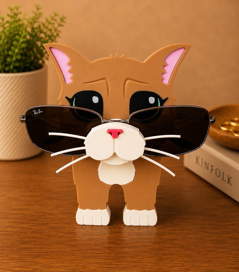 May include: A brown cat-shaped sunglasses holder with black sunglasses. The holder has pink inner ears, a white muzzle, and white whiskers. The sunglasses have a silver frame and dark lenses. The cat is standing on a wooden surface.