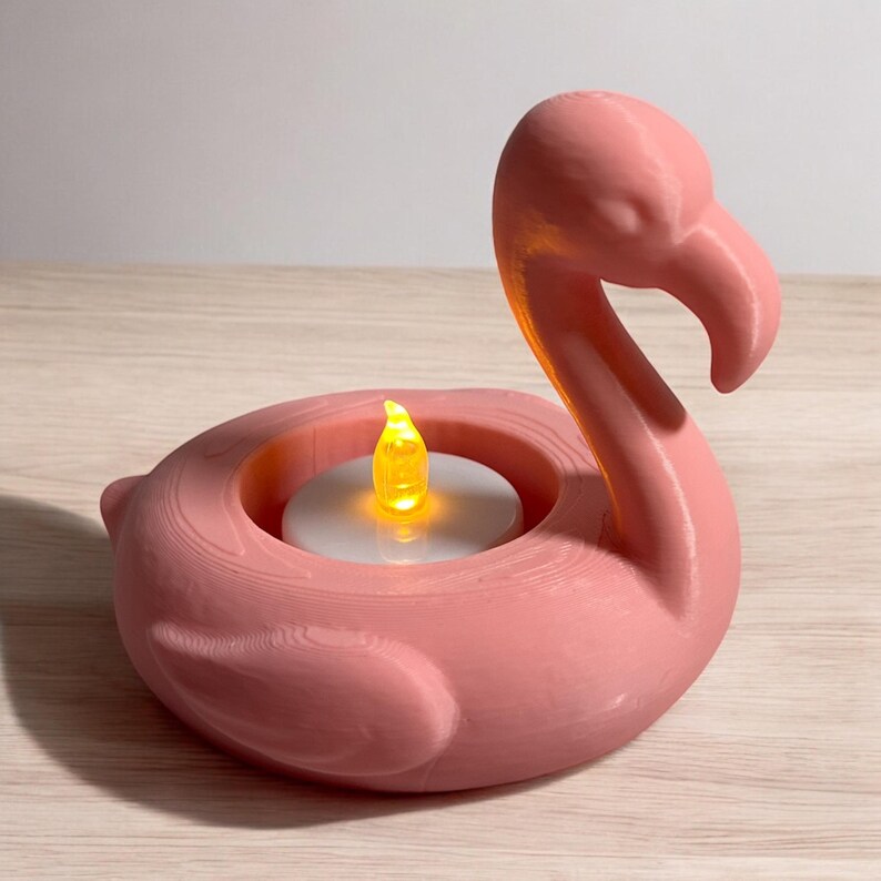 May include: A pink flamingo-shaped candle holder with a glowing LED candle. The flamingo has a long neck and beak, and the candle sits in the center of the flamingo's body. The candle holder is made of a solid material and is sitting on a light-colored surface.