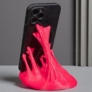 Peut inclure: Un smartphone noir est maintenu par un support de téléphone rose vif. Le support a un design unique et abstrait avec des formes fluides et fondues. Le support est fait d'un matériau solide et est conçu pour maintenir un téléphone en position verticale.
