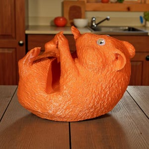 K&ouml;nnte beinhalten: Ein orangefarbener Weinflaschenhalter in Eichh&ouml;rnchenform. Die Skulptur hat eine strukturierte Oberfl&auml;che und ist mit erhobenen Pfoten dargestellt. Der Halter ist so konzipiert, dass er eine Weinflasche aufnehmen kann. Der Hintergrund zeigt eine K&uuml;che.