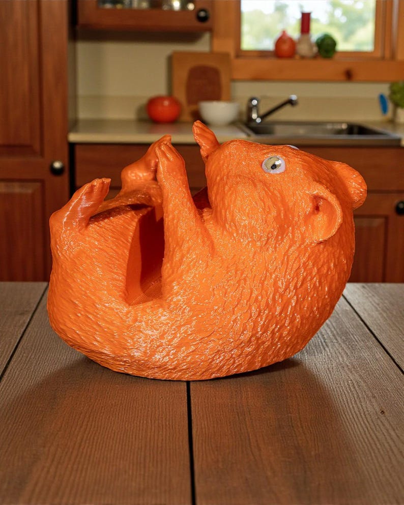K&ouml;nnte beinhalten: Ein orangefarbener Weinflaschenhalter in Eichh&ouml;rnchenform. Die Skulptur hat eine strukturierte Oberfl&auml;che und ist mit erhobenen Pfoten dargestellt. Der Halter ist so konzipiert, dass er eine Weinflasche aufnehmen kann. Der Hintergrund zeigt eine K&uuml;che.