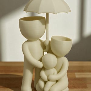 May include: A cream-colored family figurine, featuring a parent figure holding an umbrella over a family of three. The figures have open-top heads, possibly for use as planters or decorative items. The design is simple and modern.