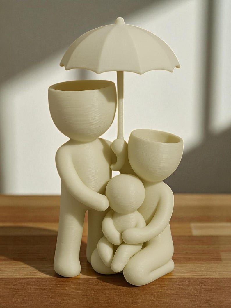 May include: A cream-colored family figurine, featuring a parent figure holding an umbrella over a family of three. The figures have open-top heads, possibly for use as planters or decorative items. The design is simple and modern.
