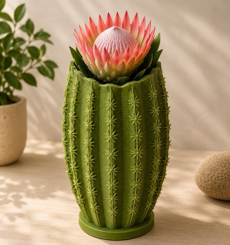 May include: A green cactus-shaped planter holds a large protea flower with pink and cream petals. The planter has a textured surface with star-shaped details, and the flower's center is a soft pink. The planter is on a light-colored surface.