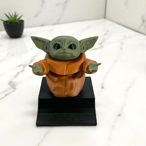 May include: A green-skinned character figurine with large ears and dark eyes, wearing an orange robe. The figure is on a black rectangular base. A small potted plant is in the background.