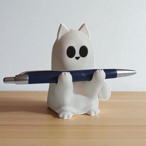 May include: A white cat-shaped pen holder with black eyes and a small black nose, holding a dark blue pen. The pen holder is sitting on a wooden surface.