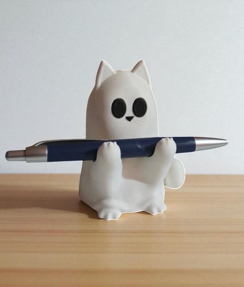 May include: A white cat-shaped pen holder with black eyes and a small black nose, holding a dark blue pen. The pen holder is sitting on a wooden surface.
