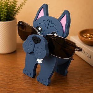 May include: A navy blue bulldog-shaped eyeglass holder with pink inner ears and a white star on its chest. The holder is wearing sunglasses with dark lenses. A small potted plant and a wooden bowl are in the background.