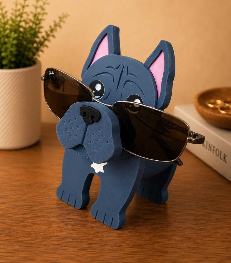 May include: A navy blue bulldog-shaped eyeglass holder with pink inner ears and a white star on its chest. The holder is wearing sunglasses with dark lenses. A small potted plant and a wooden bowl are in the background.