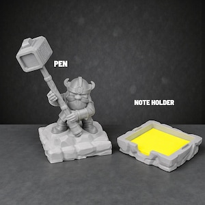 May include: A grey pen holder in the shape of a dwarf with a hammer, and a matching note holder with yellow sticky notes. The pen holder has the word "PEN" printed on it, and the note holder has the words "NOTE HOLDER" printed on it.