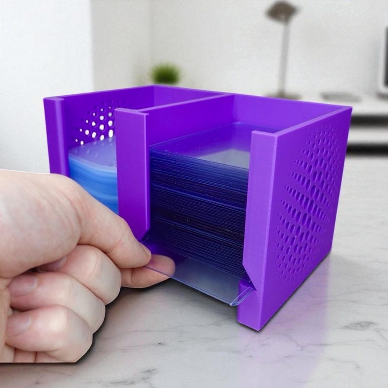 May include: A purple card holder with two compartments. One compartment holds a stack of blue cards, and the other holds a stack of clear card sleeves. The holder has a geometric pattern on the side.