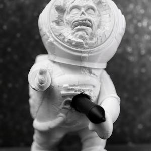 Pu&ograve; includere: Una statuetta bianca di un astronauta con un volto dettagliato all'interno del casco. L'astronauta tiene in mano un oggetto nero. La statuetta &egrave; su una superficie.