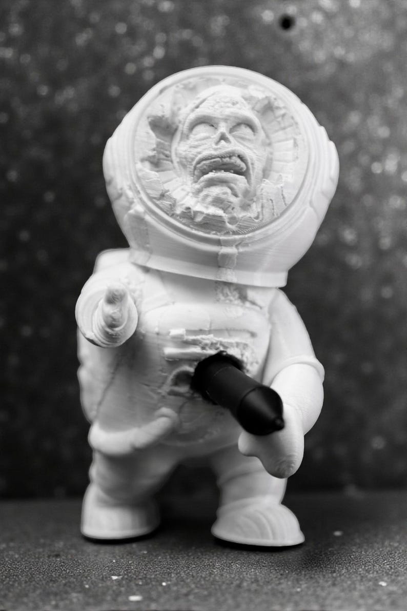 Pu&ograve; includere: Una statuetta bianca di un astronauta con un volto dettagliato all'interno del casco. L'astronauta tiene in mano un oggetto nero. La statuetta &egrave; su una superficie.