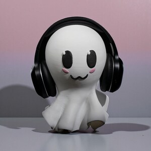 May include: A white ghost figurine with black headphones, black eyes, and pink cheeks. The ghost has a smiling expression and a draped sheet-like body. The background is a gradient of pink and purple.
