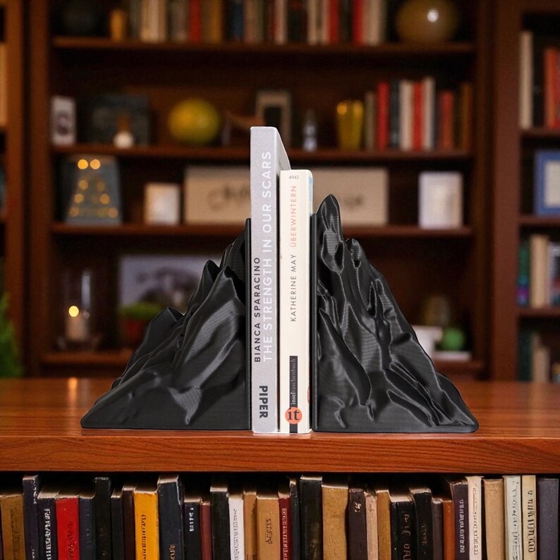 May include: Black mountain-shaped bookends holding two books. The bookends have a textured surface and are made of a solid material. The books are titled "The Strength in Our Scars" and "&Uuml;berwintern". The bookends are on a wooden surface.