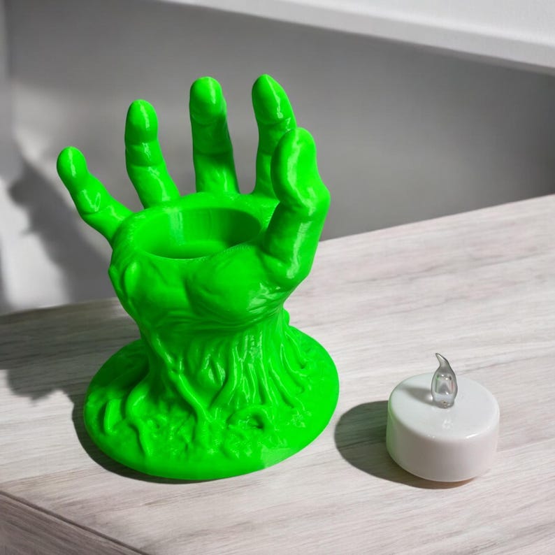 May include: A bright green, hand-shaped candle holder with a round base and a central cavity for a candle. A small, white LED candle sits beside the holder. The holder has a textured, organic design, perfect for Halloween or spooky decor.