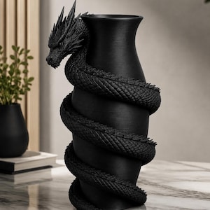 May include: A black vase with a dragon wrapped around it. The dragon's head is at the top, and its body coils around the vase. The vase is on a marble surface. A small black vase and books are in the background.