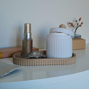 Puede incluir: Una bandeja beige de forma ovalada con borde acanalado contiene una botella pequeña, un reloj plateado y un recipiente blanco con tapa. El recipiente tiene ranuras verticales. Una pequeña rama de flores está en un jarrón en el fondo.