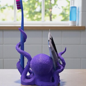 Puede incluir: Un soporte para cepillo de dientes con forma de pulpo morado con un cepillo de dientes azul y un tubo de pasta de dientes. El cepillo de dientes tiene un cabezal de cerdas rosa y morado. El soporte est&aacute; sobre una superficie de madera frente a una ventana.
