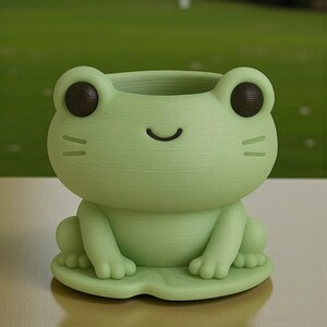 Puede incluir: Una maceta en forma de rana verde claro con una cara sonriente, ojos marrones oscuros y peque&ntilde;os bigotes. La maceta se asienta sobre una base de hoja de nen&uacute;far verde. La boca abierta de la rana forma un peque&ntilde;o recipiente.