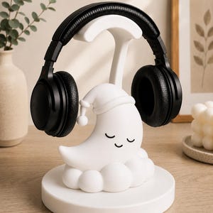 May include: A white headphone stand featuring a crescent moon design wearing a nightcap, resting on a cloud base. Black headphones are placed on the stand. The stand is on a round base.
