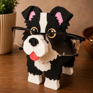 May include: A black and white dog-shaped eyeglasses holder with a white snout, red tongue, and black framed glasses. The dog has pink inner ears and is made of wood. The holder is on a wooden surface.