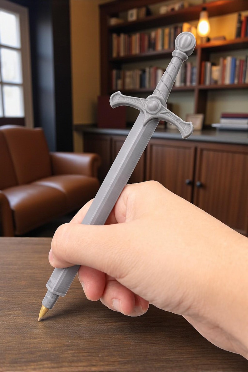May include: A gray pen shaped like a medieval sword, held in a hand. The pen has a detailed hilt and a gold-colored tip. The background includes a wooden table, a bookshelf, and a leather armchair.