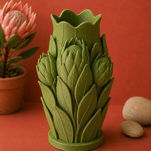 Puede incluir: Un jarr&oacute;n decorativo verde con un dise&ntilde;o floral. El jarr&oacute;n tiene forma de flor con hojas y p&eacute;talos detallados. El jarr&oacute;n est&aacute; sobre una superficie rojo-naranja, con una planta en maceta y dos piedras en el fondo.
