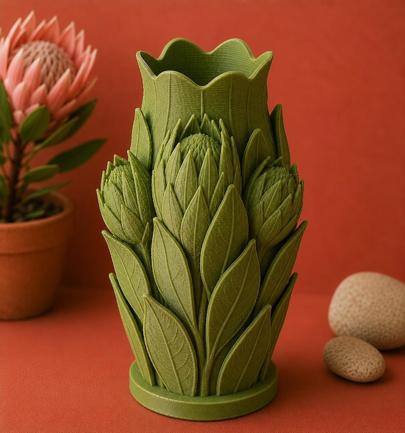 Puede incluir: Un jarr&oacute;n decorativo verde con un dise&ntilde;o floral. El jarr&oacute;n tiene forma de flor con hojas y p&eacute;talos detallados. El jarr&oacute;n est&aacute; sobre una superficie rojo-naranja, con una planta en maceta y dos piedras en el fondo.